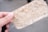 A close-up of a hand holding a textured piece of handmade paper with visible fibers and specks. The paper appears fibrous and natural, with a light beige color contrasting with the soft focus background.