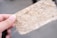 A close-up of a hand holding a textured piece of handmade paper with visible fibers and specks. The paper appears fibrous and natural, with a light beige color contrasting with the soft focus background.