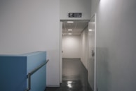 A hallway in what appears to be an office or institutional building with white walls and a dark reflective floor. The hallway is illuminated by ceiling lights and leads to a door marked with a sign for the third floor. A blue ramp with a metal handrail is visible on the left side.