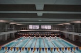 A large indoor swimming pool with multiple lanes separated by lane markers. The pool is surrounded by a concrete structure with an upper level walkway. Blue and yellow triangular banners are hung above the pool. Swimmers can be seen in some of the lanes, and there are starting blocks at one end of the pool.