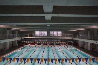 A large indoor swimming pool with multiple lanes separated by lane markers. The pool is surrounded by a concrete structure with an upper level walkway. Blue and yellow triangular banners are hung above the pool. Swimmers can be seen in some of the lanes, and there are starting blocks at one end of the pool.