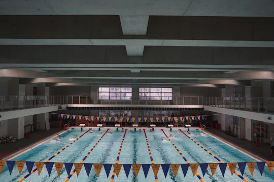 A large indoor swimming pool with multiple lanes separated by lane markers. The pool is surrounded by a concrete structure with an upper level walkway. Blue and yellow triangular banners are hung above the pool. Swimmers can be seen in some of the lanes, and there are starting blocks at one end of the pool.