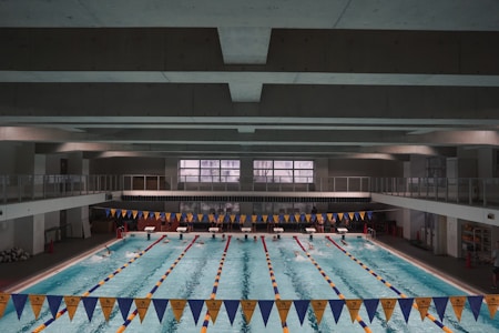 A large indoor swimming pool with multiple lanes separated by lane markers. The pool is surrounded by a concrete structure with an upper level walkway. Blue and yellow triangular banners are hung above the pool. Swimmers can be seen in some of the lanes, and there are starting blocks at one end of the pool.