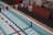 An indoor swimming pool with clear blue water and colorful lane markers. Yellow and blue triangular flags are strung above the pool. There is a large wall clock, a folded wooden panel, and some red cones near the poolside.