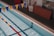 An indoor swimming pool with clear blue water and colorful lane markers. Yellow and blue triangular flags are strung above the pool. There is a large wall clock, a folded wooden panel, and some red cones near the poolside.