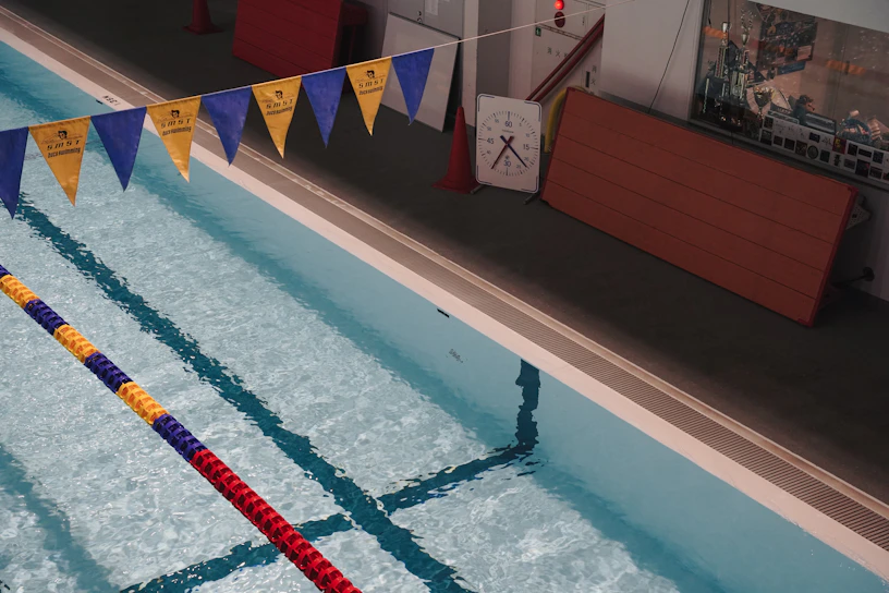 An indoor swimming pool with clear blue water and colorful lane markers. Yellow and blue triangular flags are strung above the pool. There is a large wall clock, a folded wooden panel, and some red cones near the poolside.