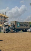 A large container truck is parked on a sandy surface with a Maersk shipping container mounted on it. The background shows a multi-story building with yellow and white coloring and several parked cars. The sky is partially cloudy, adding a serene atmosphere.
