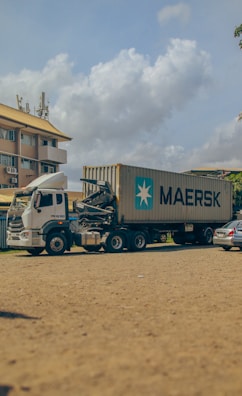 A large container truck is parked on a sandy surface with a Maersk shipping container mounted on it. The background shows a multi-story building with yellow and white coloring and several parked cars. The sky is partially cloudy, adding a serene atmosphere.