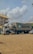 A large container truck is parked on a sandy surface with a Maersk shipping container mounted on it. The background shows a multi-story building with yellow and white coloring and several parked cars. The sky is partially cloudy, adding a serene atmosphere.