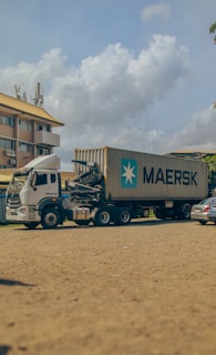 A large container truck is parked on a sandy surface with a Maersk shipping container mounted on it. The background shows a multi-story building with yellow and white coloring and several parked cars. The sky is partially cloudy, adding a serene atmosphere.