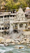 A riverside temple with intricate architecture is set against a lush, green hillside. Steam rises from rocks near the river, indicating geothermal activity. Colorful prayer flags hang across the scene, adding vibrancy.