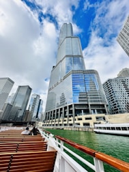 A striking skyscraper with reflective glass windows towers upwards under a partially cloudy sky. The building is surrounded by other tall buildings in an urban setting. In the foreground, there is a river with greenish water and an empty seating area on a boat, featuring wooden benches and white railings.