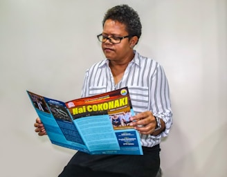 A person with short curly hair and glasses is seated while reading a colorful magazine. The magazine has a red and yellow title with multiple sections and images. The person is wearing a white and black striped shirt and has a focused expression.