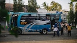 A blue and black bus with the letters 'BSK' on its side is parked on a street. Several people stand nearby, some engaging in conversation, while others appear to be boarding or disembarking the bus. The background shows trees and a few buildings, including a visible BWS sign.
