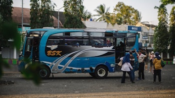 A blue and black bus with the letters 'BSK' on its side is parked on a street. Several people stand nearby, some engaging in conversation, while others appear to be boarding or disembarking the bus. The background shows trees and a few buildings, including a visible BWS sign.