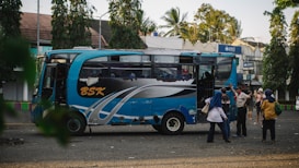 A blue and black bus with the letters 'BSK' on its side is parked on a street. Several people stand nearby, some engaging in conversation, while others appear to be boarding or disembarking the bus. The background shows trees and a few buildings, including a visible BWS sign.