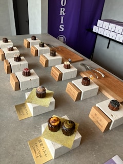 A display of various pastries placed on white stone blocks on a counter. Each pastry is labeled with a golden or wooden sign. There are utensils like tongs and knives placed next to the pastries on wooden boards. In the background, there is a purple banner with text and a shelf with stacked white boxes.