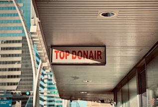 A sign reading 'Top Donair' hangs under a building canopy in an urban area, with modern high-rise buildings visible in the background.