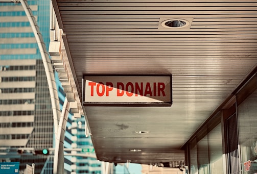 A sign reading 'Top Donair' hangs under a building canopy in an urban area, with modern high-rise buildings visible in the background.