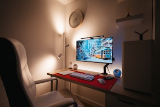 A modern, minimalistic workspace featuring a large monitor displaying a vibrant cityscape with a tram. A red mouse pad rests on the desk, accompanied by a keyboard, mouse, and a small speaker. A white chair complements the neutral-toned walls and soft lighting. A circular wall clock and ambient lighting add to the aesthetic and cozy setup.