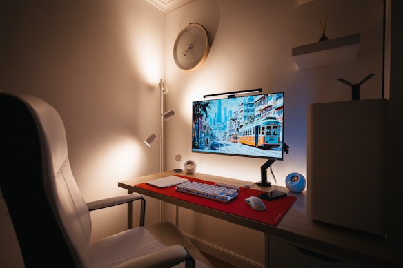 A modern, minimalistic workspace featuring a large monitor displaying a vibrant cityscape with a tram. A red mouse pad rests on the desk, accompanied by a keyboard, mouse, and a small speaker. A white chair complements the neutral-toned walls and soft lighting. A circular wall clock and ambient lighting add to the aesthetic and cozy setup.