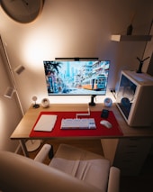 A clean and organized computer workspace features a large monitor displaying a cityscape with trams, placed on a wooden desk with a red mat. The setup includes a white mechanical keyboard and a wireless mouse, alongside a closed laptop. Surrounding the monitor are small speakers and a smartphone, with a white PC tower on the right. The ambient lighting creates a warm and cozy atmosphere, complemented by a wall clock, a shelf with a vase, and a stylish chair.