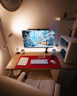 A clean and organized computer workspace features a large monitor displaying a cityscape with trams, placed on a wooden desk with a red mat. The setup includes a white mechanical keyboard and a wireless mouse, alongside a closed laptop. Surrounding the monitor are small speakers and a smartphone, with a white PC tower on the right. The ambient lighting creates a warm and cozy atmosphere, complemented by a wall clock, a shelf with a vase, and a stylish chair.