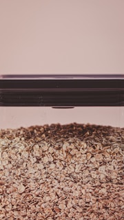 A large, clear glass container filled with a substantial quantity of oat flakes. The lid of the container is dark and tightly sealed. The background is a soft beige color, giving the image a warm and neutral tone.