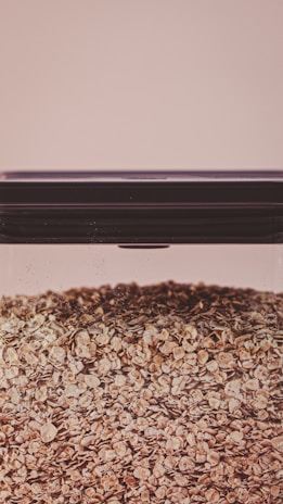 A large, clear glass container filled with a substantial quantity of oat flakes. The lid of the container is dark and tightly sealed. The background is a soft beige color, giving the image a warm and neutral tone.