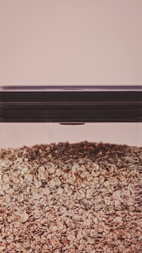 A large, clear glass container filled with a substantial quantity of oat flakes. The lid of the container is dark and tightly sealed. The background is a soft beige color, giving the image a warm and neutral tone.