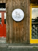 A wooden wall features a circular sign depicting a sheep with the text 'Icelandic Street Food'. The wall has a rustic appearance with vertical planks. On the left, there is a red door, and to the right, a window with a yellow frame displaying patterns of ice cream cones.