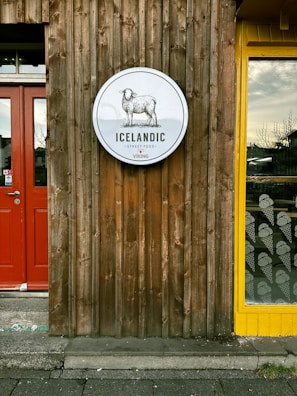 A wooden wall features a circular sign depicting a sheep with the text 'Icelandic Street Food'. The wall has a rustic appearance with vertical planks. On the left, there is a red door, and to the right, a window with a yellow frame displaying patterns of ice cream cones.