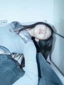 A person is reclining on a surface with closed eyes, appearing to be in a relaxed state. The individual is wearing a light-colored top and gray jeans. The setting appears minimalistic and dimly lit, creating a serene and calm atmosphere.
