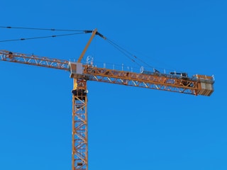 A tall construction crane with a long horizontal arm extends against a clear blue sky. The crane is yellow and consists of metal lattice structures, showing pulleys and cables used for lifting.