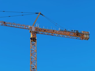 A tall construction crane with a long horizontal arm extends against a clear blue sky. The crane is yellow and consists of metal lattice structures, showing pulleys and cables used for lifting.