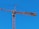 A tall construction crane with a long horizontal arm extends against a clear blue sky. The crane is yellow and consists of metal lattice structures, showing pulleys and cables used for lifting.