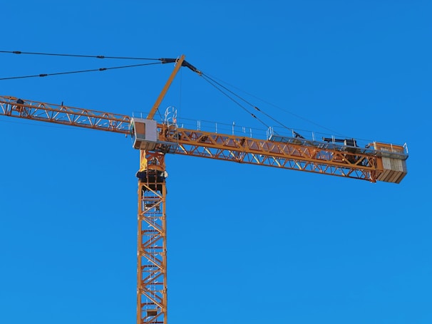 A tall construction crane with a long horizontal arm extends against a clear blue sky. The crane is yellow and consists of metal lattice structures, showing pulleys and cables used for lifting.