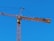 A tall construction crane with a long horizontal arm extends against a clear blue sky. The crane is yellow and consists of metal lattice structures, showing pulleys and cables used for lifting.