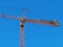 A tall construction crane with a long horizontal arm extends against a clear blue sky. The crane is yellow and consists of metal lattice structures, showing pulleys and cables used for lifting.