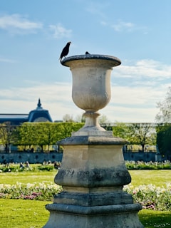 A large stone urn is prominently positioned in a lush garden setting. A bird is perched on the rim of the urn, with a clear sky and the silhouette of a building visible in the background. The garden is filled with green grass and colorful flowers.