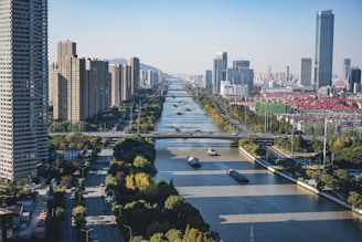 A modern urban landscape featuring a wide river running through a city with numerous skyscrapers and tall buildings on either side. Several barges are seen navigating the river. The cityscape includes a mix of greenery and urban development, with clear blue skies and distant mountains in the background.