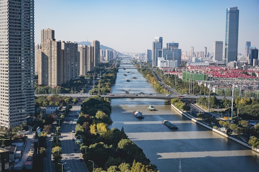 A modern urban landscape featuring a wide river running through a city with numerous skyscrapers and tall buildings on either side. Several barges are seen navigating the river. The cityscape includes a mix of greenery and urban development, with clear blue skies and distant mountains in the background.