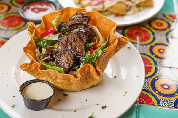 A tortilla shell forms a bowl holding a vibrant salad with mixed greens, red bell peppers, and grilled slices of steak. Surrounding the dish, there's a small container of creamy dressing on the plate. The table has colorful, patterned tiles with geometric designs. A separate small bowl of red sauce rests nearby.