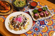 A vibrant tabletop adorned with colorful tiles hosting a variety of Mexican dishes. In the center is a plate of nachos topped with black beans, chopped tomatoes, shredded lettuce, sour cream, and pickled red onions. To the side, there is a serving tray with three tacos filled with meat, garnished with pink onions and slices of jalapeño. Flanking these dishes are bowls of guacamole, red, and green salsa.