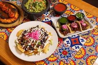 A vibrant tabletop adorned with colorful tiles hosting a variety of Mexican dishes. In the center is a plate of nachos topped with black beans, chopped tomatoes, shredded lettuce, sour cream, and pickled red onions. To the side, there is a serving tray with three tacos filled with meat, garnished with pink onions and slices of jalape&ntilde;o. Flanking these dishes are bowls of guacamole, red, and green salsa.