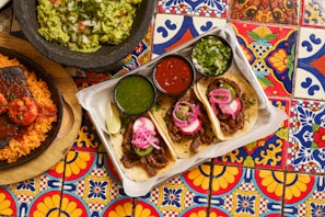 A vibrant and colorful table setting with a platter of three tacos topped with pickled onions and cilantro, accompanied by lime wedges and small bowls of green sauce, red salsa, and additional cilantro. To the side, there is a stone bowl of guacamole with chopped onions and tomatoes, and a plate of rice and grilled meat with sauce. The table is adorned with intricate, multicolored tiles featuring floral and geometric patterns.