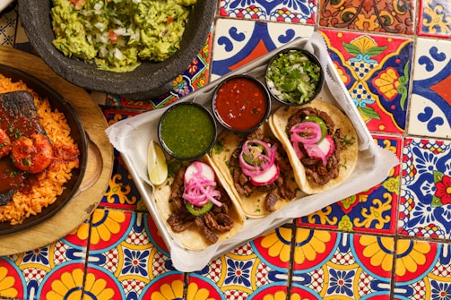 A vibrant and colorful table setting with a platter of three tacos topped with pickled onions and cilantro, accompanied by lime wedges and small bowls of green sauce, red salsa, and additional cilantro. To the side, there is a stone bowl of guacamole with chopped onions and tomatoes, and a plate of rice and grilled meat with sauce. The table is adorned with intricate, multicolored tiles featuring floral and geometric patterns.