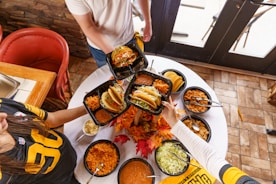 A group of people is gathered around a table laden with Mexican cuisine. The table is adorned with bowls of vibrant rice, tacos, shredded lettuce, various salsas, and a decorative centerpiece with autumn leaves. Two individuals holding plates filled with tacos are visible, wearing sports jerseys, possibly suggesting a tailgate or casual gathering.