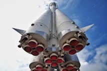 A large rocket viewed from below, showing its multiple red exhaust nozzles and metallic silver body, against a backdrop of blue sky with some clouds.