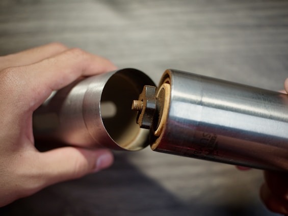 A pair of hands is holding a metallic cylindrical object that appears to be a socket wrench or a similar tool. The object is shiny with a smooth surface, and has a nut or bolt partially inserted into it, demonstrating some mechanical function or assembly process. The background is blurred and features a surface that might be wood.